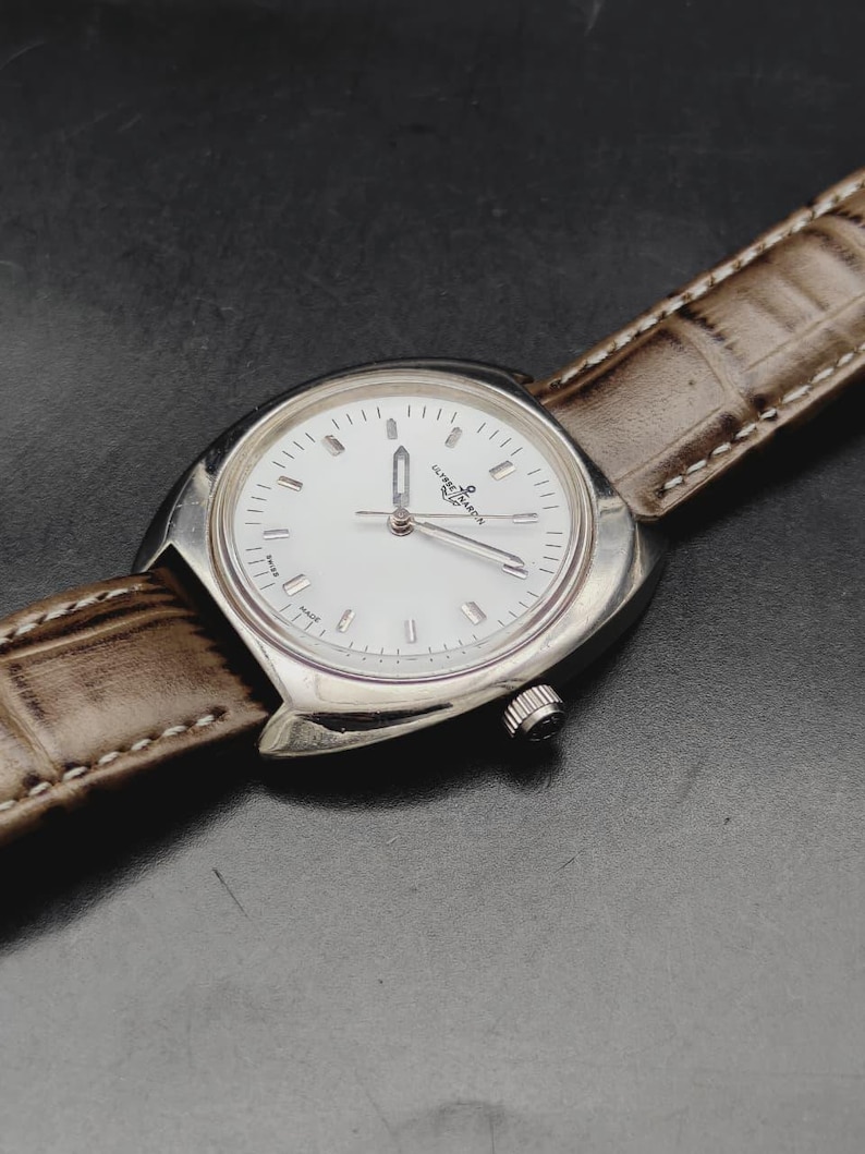 May include: A silver-toned wristwatch with a white dial, silver hour markers, and silver hands. The watch features the Ulysse Nardin logo on the face and is attached to a textured, light brown leather strap with visible white stitching along the edges.