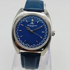 Puede incluir: Un reloj Jaeger-LeCoultre Club vintage con esfera azul y caja de acero inoxidable. El reloj tiene una correa de cuero azul y una corona plateada. La esfera tiene marcadores de hora plateados y el logotipo de Jaeger-LeCoultre.