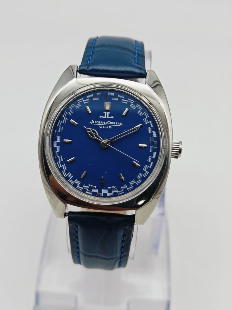 Puede incluir: Un reloj Jaeger-LeCoultre Club vintage con esfera azul y caja de acero inoxidable. El reloj tiene una correa de cuero azul y una corona plateada. La esfera tiene marcadores de hora plateados y el logotipo de Jaeger-LeCoultre.
