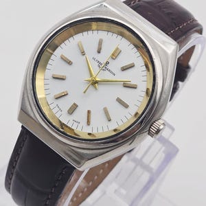 Pu&ograve; includere: Un orologio Ulysse Nardin argentato con quadrante bianco, dettagli dorati e cinturino in pelle marrone. L'orologio ha una cassa rettangolare e un design classico. Il quadrante mostra il nome del marchio e la scritta "SWISS MADE".
