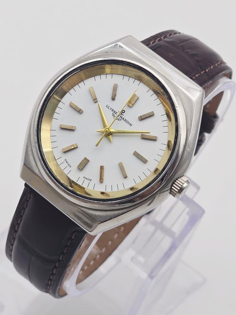 Pu&ograve; includere: Un orologio Ulysse Nardin argentato con quadrante bianco, dettagli dorati e cinturino in pelle marrone. L'orologio ha una cassa rettangolare e un design classico. Il quadrante mostra il nome del marchio e la scritta "SWISS MADE".