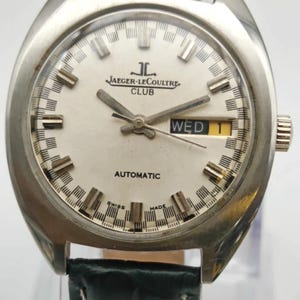 Puede incluir: Un reloj autom&aacute;tico Jaeger-LeCoultre Club vintage con caja plateada y esfera blanca. La esfera muestra el nombre de la marca, d&iacute;a y fecha, y agujas luminosas. El reloj tiene una correa de cuero verde.
