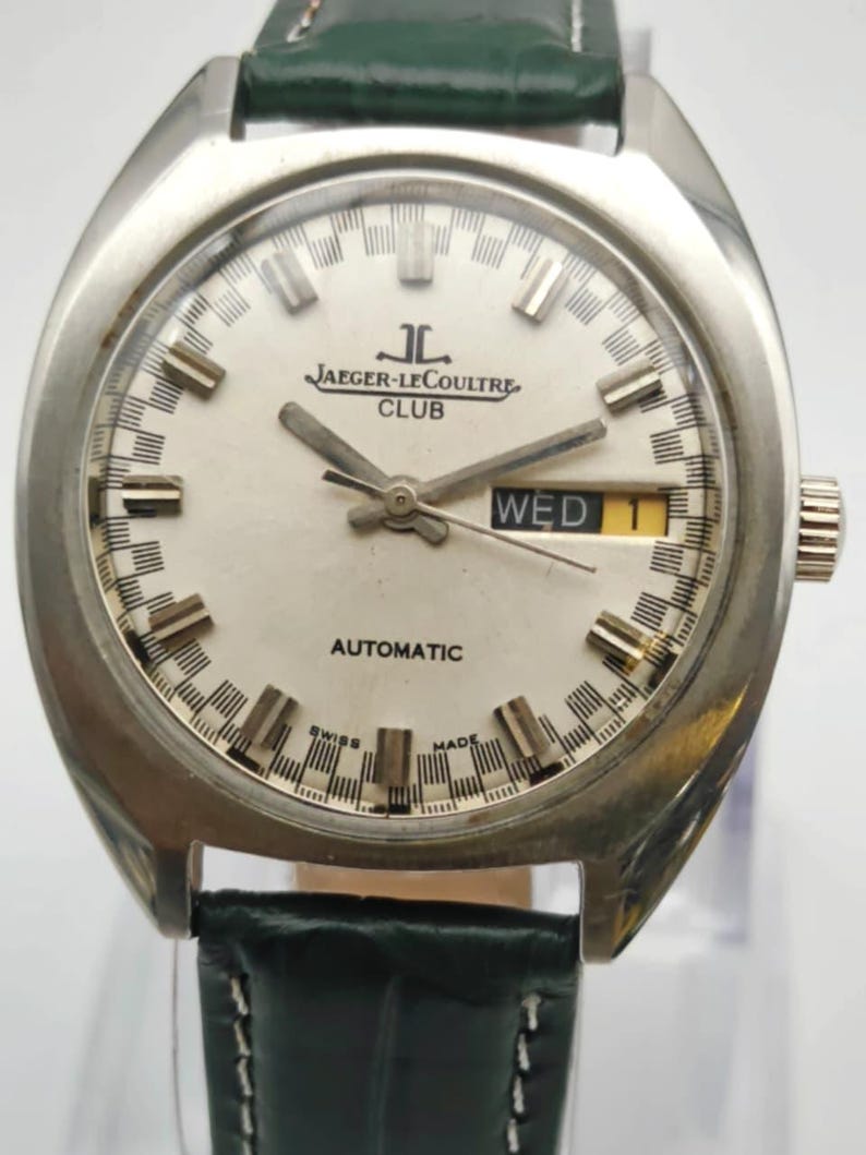 Puede incluir: Un reloj autom&aacute;tico Jaeger-LeCoultre Club vintage con caja plateada y esfera blanca. La esfera muestra el nombre de la marca, d&iacute;a y fecha, y agujas luminosas. El reloj tiene una correa de cuero verde.