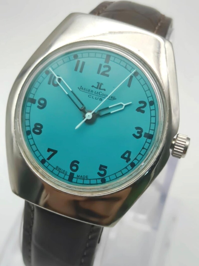 Op de afbeelding: Een zilverkleurig Jaeger-LeCoultre Club horloge met een turquoise wijzerplaat en zwarte cijfers. Het horloge heeft een bruine leren band en een zilveren lunette. De wijzerplaat toont het Jaeger-LeCoultre Club logo en de tekst "SWISS MADE".