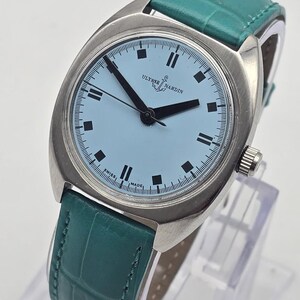 Peut inclure: Une montre-bracelet argent&eacute;e avec un cadran bleu clair et des aiguilles noires. La montre porte l'inscription "ULYSSE NARDIN" et un bracelet en cuir vert. Le cadran comporte des index noirs rectangulaires et une trotteuse.