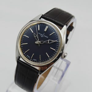 May include: A silver-toned Ulysse Nardin wristwatch with a dark blue face and black leather band. The watch features silver hands and markers, and the words "Ulysse Nardin" are printed on the face. The band has a crocodile pattern.