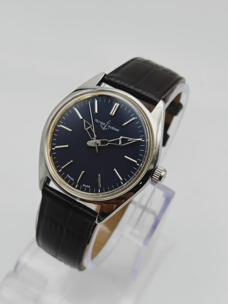 May include: A silver-toned Ulysse Nardin wristwatch with a dark blue face and black leather band. The watch features silver hands and markers, and the words "Ulysse Nardin" are printed on the face. The band has a crocodile pattern.