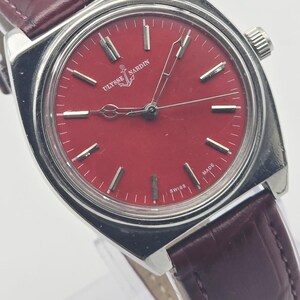 May include: A vintage Ulysse Nardin wristwatch with a red dial and silver-tone case. The watch features a burgundy leather strap and the brand's logo. The dial has silver markers and hands. The watch is marked "SWISS MADE".
