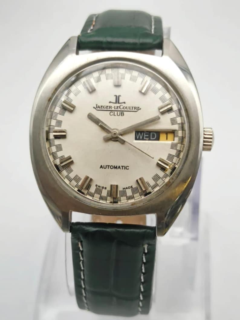 Puede incluir: Un reloj Jaeger-LeCoultre Club vintage con caja plateada y esfera blanca. El reloj presenta una pantalla de d&iacute;a y fecha y una correa de cuero verde. La esfera muestra el texto "AUTOMATIC" y "SWISS MADE".