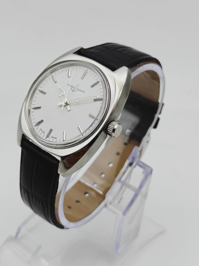 May include: A silver-toned wristwatch with a white face and black leather band. The watch features silver hands and hour markers. The band is textured. The watch is displayed on a clear stand.