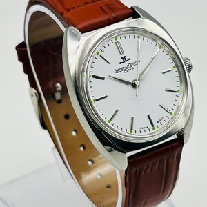 May include: A silver-toned Jaeger-LeCoultre Club watch with a white face and brown leather band. The watch features a round face with black and green markers and the Jaeger-LeCoultre Club logo. The brown leather band has a textured pattern.