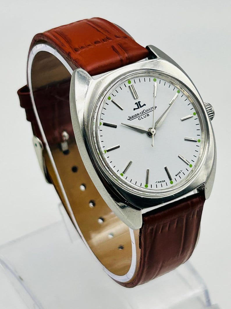 May include: A silver-toned Jaeger-LeCoultre Club watch with a white face and brown leather band. The watch features a round face with black and green markers and the Jaeger-LeCoultre Club logo. The brown leather band has a textured pattern.