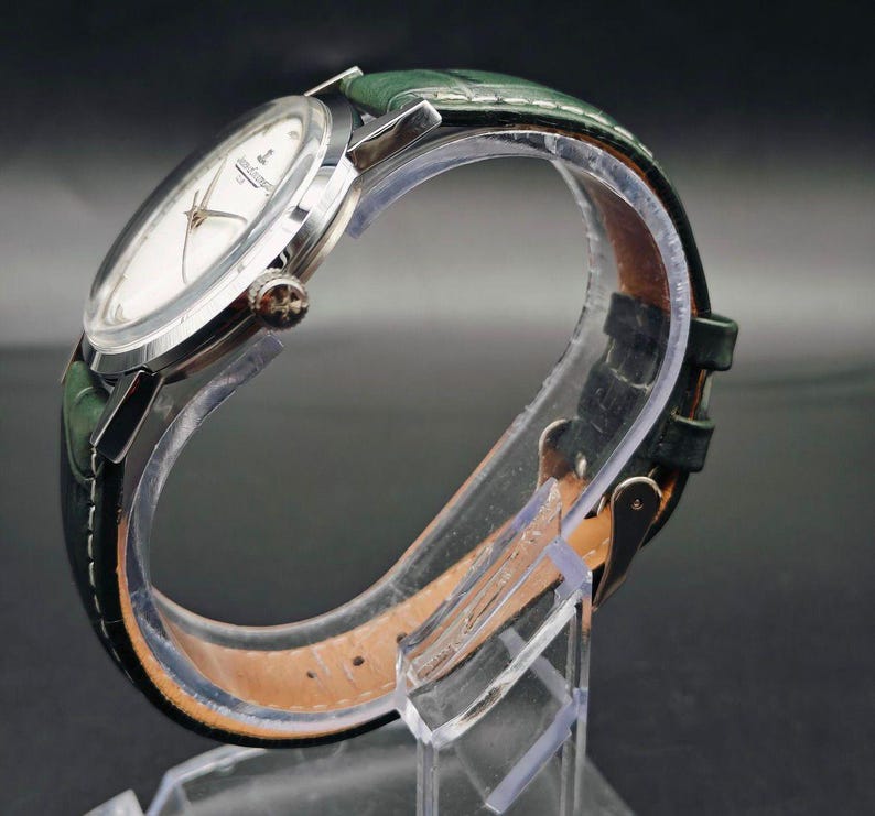 Pu&ograve; includere: Un orologio da polso argentato con quadrante bianco e cinturino in pelle verde. L'orologio ha un quadrante rotondo con lancette e indici argentati. Il cinturino verde &egrave; strutturato e ha una chiusura a fibbia. L'orologio &egrave; esposto su un supporto trasparente.