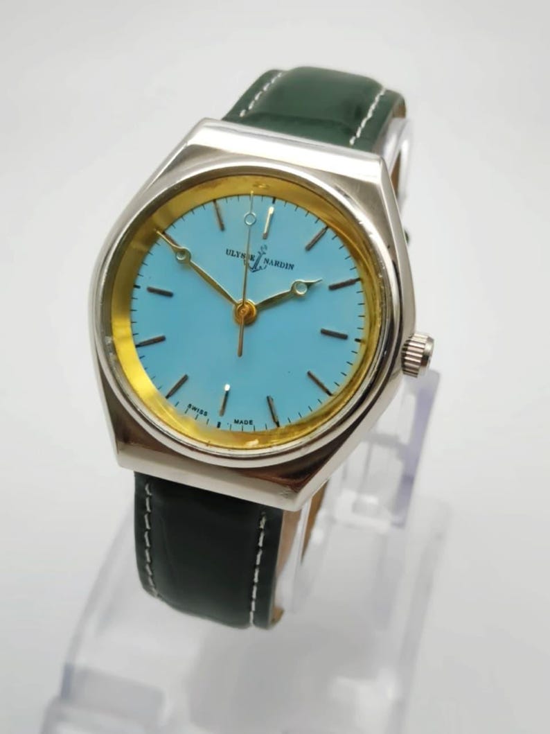 May include: A silver-toned wristwatch with a light blue face and gold-colored hands and accents. The watch has a green leather band with white stitching. The watch face has the text "ULYSSES NARDIN SWISS MADE".