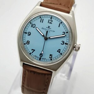 K&ouml;nnte beinhalten: Eine silberfarbene Armbanduhr mit hellblauem Zifferblatt und braunem Lederarmband. Das Zifferblatt zeigt schwarze Ziffern und Zeiger, mit dem Markennamen "Jaeger-LeCoultre". Ein klassisches Uhrendesign.