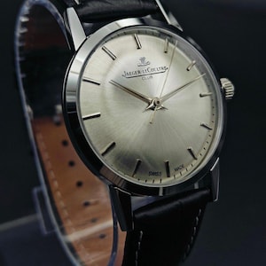 Op de afbeelding: Een zilverkleurig Jaeger-LeCoultre Club horloge met een zwarte leren band. De wijzerplaat heeft een radiale geborstelde afwerking en zilveren wijzers. De band is gestikt met witte draad. Het horloge is gemarkeerd met "SWISS MADE".