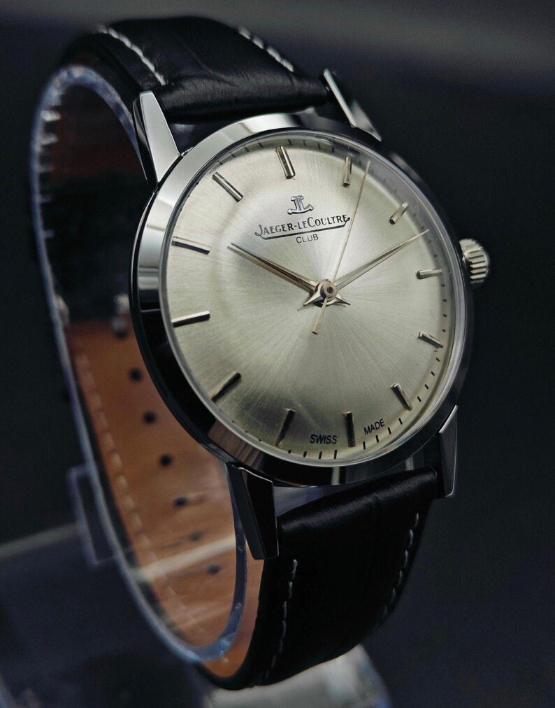 Op de afbeelding: Een zilverkleurig Jaeger-LeCoultre Club horloge met een zwarte leren band. De wijzerplaat heeft een radiale geborstelde afwerking en zilveren wijzers. De band is gestikt met witte draad. Het horloge is gemarkeerd met "SWISS MADE".