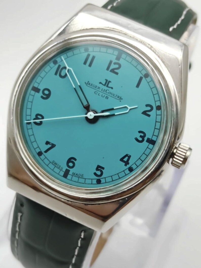 Pode incluir: Um rel&oacute;gio de pulso prateado com mostrador turquesa e algarismos pretos. O rel&oacute;gio apresenta o texto "Jaeger-LeCoultre Club" e tem uma bracelete de couro verde escuro. Os ponteiros do rel&oacute;gio s&atilde;o prateados.