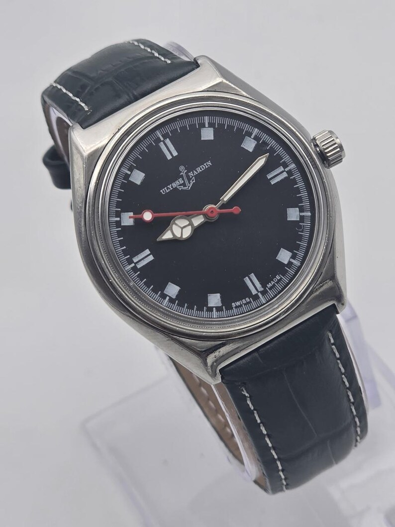 Puede incluir: Un reloj Ulysse Nardin de acero inoxidable con esfera negra y correa de cuero negro. El reloj tiene marcadores de hora blancos, un segundero rojo y una corona a la derecha. La correa mide unos 20 cm.