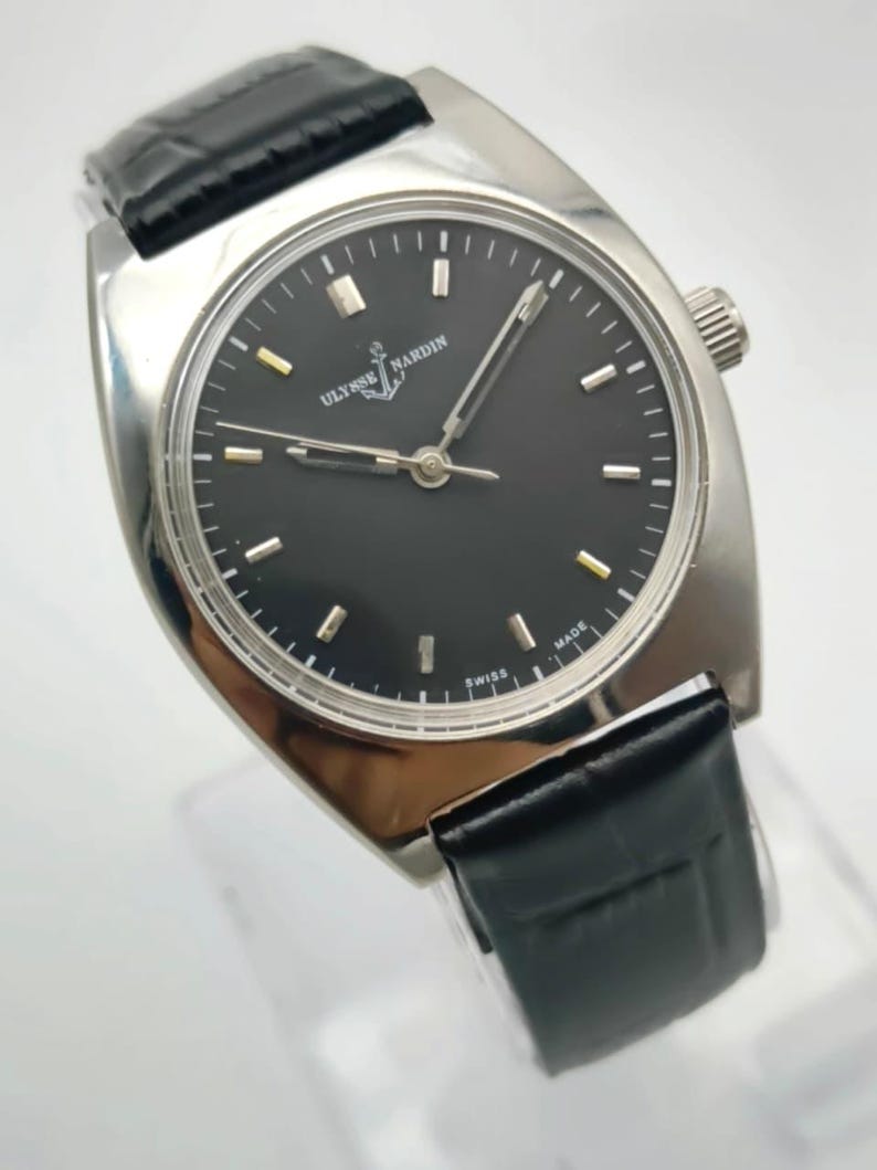Puede incluir: Un reloj de pulsera Ulysse Nardin plateado con esfera negra y correa de cuero negro. La esfera del reloj tiene marcadores y agujas plateadas, con el nombre de la marca y el logotipo de la ancla. El reloj est&aacute; marcado "SWISS MADE".