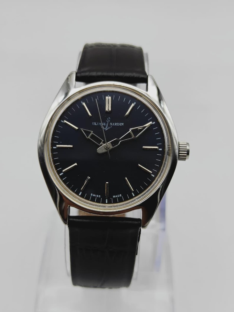 May include: A Ulysse Nardin wristwatch with a black dial and silver hands and markers. The watch has a silver case and a black leather band. The watch is displayed on a clear stand.