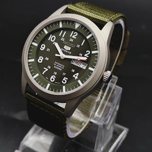 Op de afbeelding: Een Seiko 5 Sports automatisch horloge met een matte zilverkleurige kast en een olijfgroene wijzerplaat. De wijzerplaat heeft witte Arabische cijfers, een dag-datumvenster op de drie-uurpositie en een bijpassende olijfgroene stoffen band.