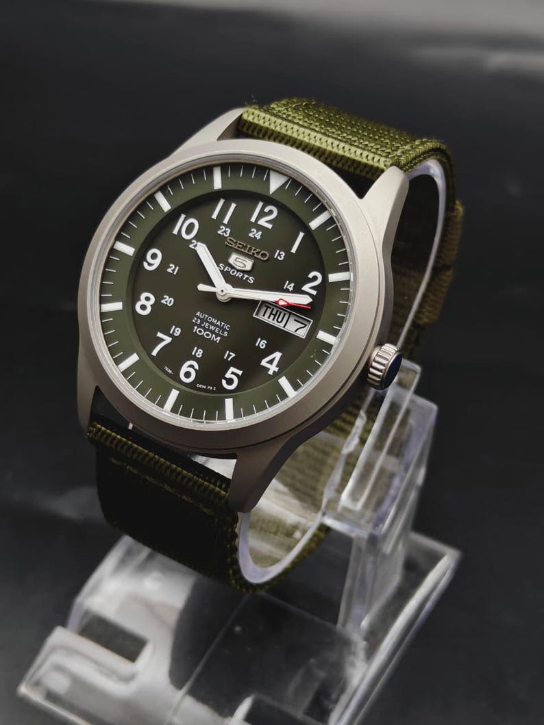 Puede incluir: Un reloj autom&aacute;tico Seiko 5 Sports con caja plateada mate y esfera verde oliva. La esfera presenta n&uacute;meros ar&aacute;bigos blancos, una ventana de d&iacute;a y fecha en la posici&oacute;n de las tres y una correa de tela verde oliva a juego con hebilla.