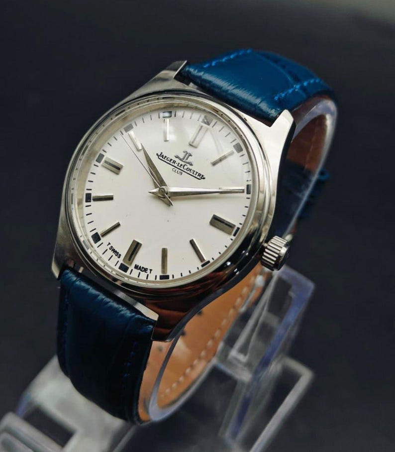 Peut inclure: Une montre Jaeger-LeCoultre Club argent&eacute;e avec un cadran blanc et un bracelet en cuir bleu. Le cadran de la montre comporte des marqueurs et des aiguilles noirs, avec le nom de la marque et "CLUB" imprim&eacute; sur le cadran. Le bracelet en cuir bleu est cousu.