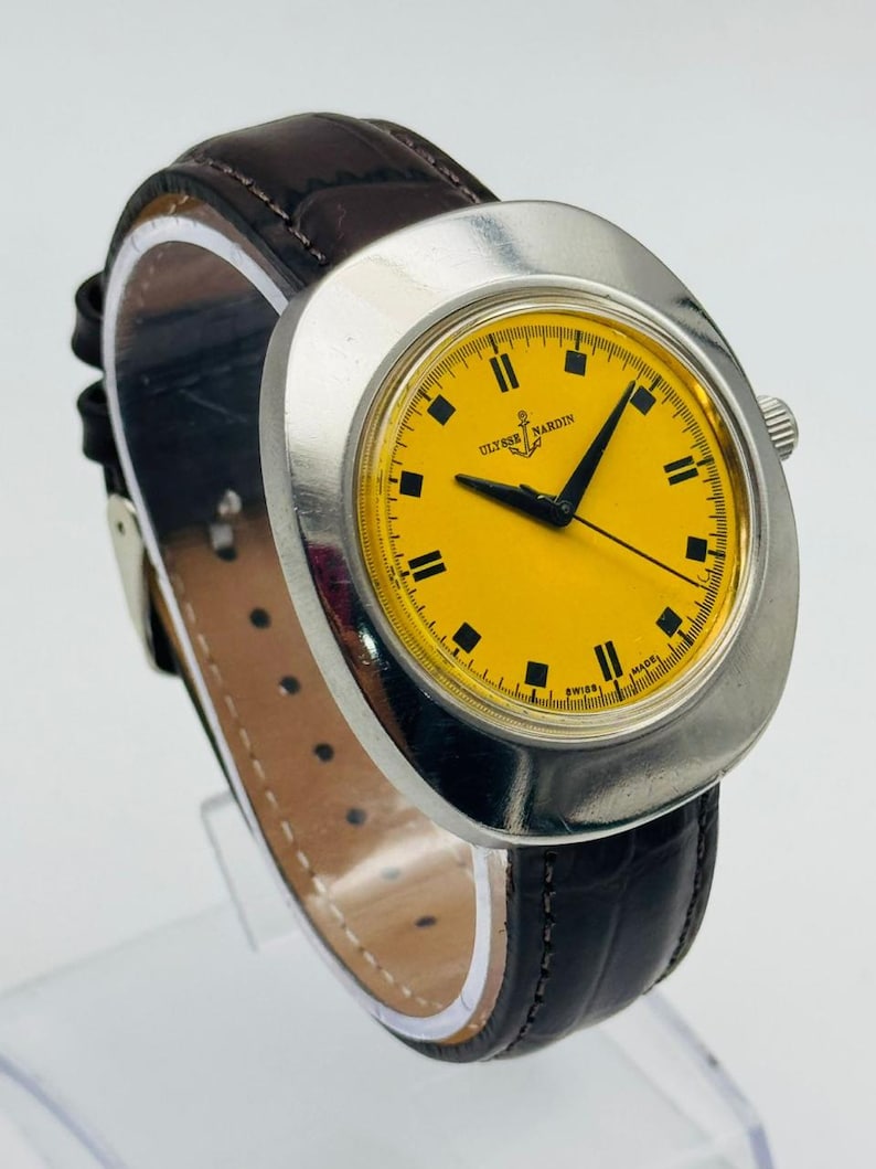 May include: A vintage Ulysse Nardin wristwatch with a bright yellow face and black markers. The watch has a silver-tone case and a dark brown leather strap. The watch face displays the brand name and an anchor logo.
