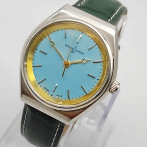 May include: A silver-toned Ulysse Nardin watch with a light blue face and gold-colored accents. The watch has a green leather strap with white stitching. The watch face displays the text "Ulysse Nardin" and "Swiss Made".