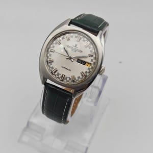 Puede incluir: Un reloj de pulsera plateado con esfera blanca, manecillas negras y visualizaci&oacute;n de d&iacute;a y fecha. El reloj tiene una correa de cuero verde. La palabra "AUTOMATIC" es visible en la esfera. El reloj se exhibe sobre un soporte transparente.