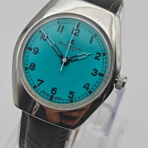Op de afbeelding: Een zilverkleurig Jaeger-LeCoultre Club horloge met een turquoise wijzerplaat en zwarte wijzers. Het horloge heeft een zwarte leren band en een zilveren kroon. De wijzerplaat toont de cijfers 1 tot en met 12. De woorden "SWISS MADE" staan op de wijzerplaat.