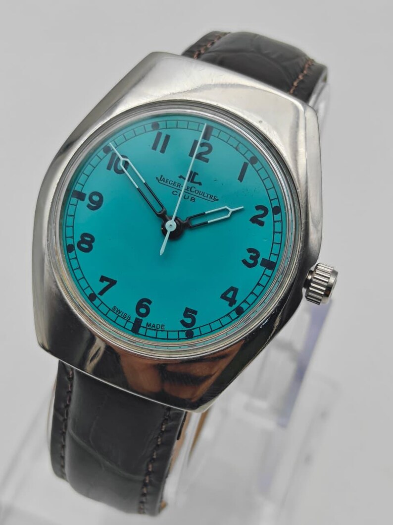 Op de afbeelding: Een zilverkleurig Jaeger-LeCoultre Club horloge met een turquoise wijzerplaat en zwarte wijzers. Het horloge heeft een zwarte leren band en een zilveren kroon. De wijzerplaat toont de cijfers 1 tot en met 12. De woorden "SWISS MADE" staan op de wijzerplaat.