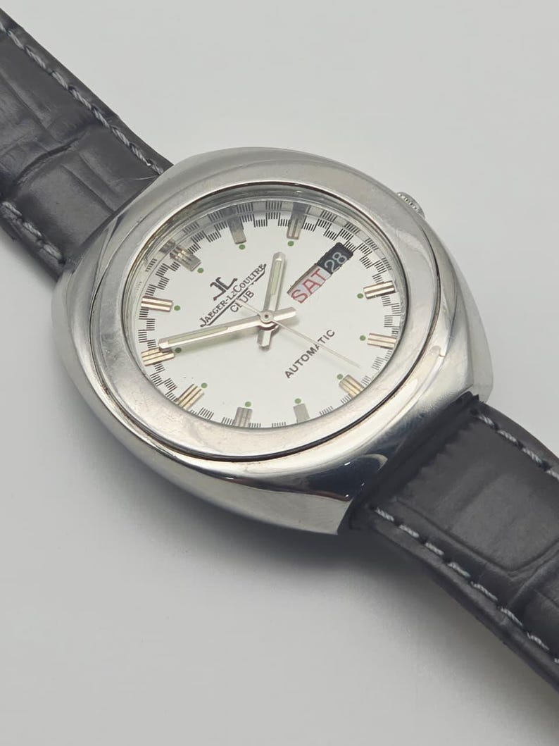 Puede incluir: Un reloj autom&aacute;tico Jaeger-LeCoultre Club plateado con esfera blanca y correa de cuero negro. La esfera muestra el d&iacute;a y la fecha, con "SAT 28" visible. El reloj tiene un dise&ntilde;o cl&aacute;sico y elegante.