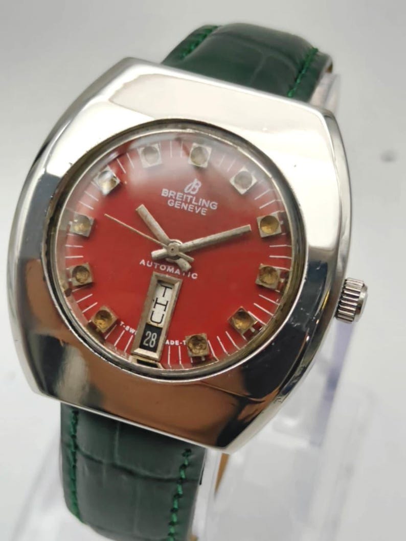 May include: A vintage Breitling Geneve automatic watch with a red dial and a green leather strap. The watch has a silver-colored case and a date display at the 6 o'clock position. The watch face has gold-colored hour markers.