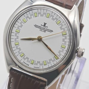 May include: A silver-toned Jaeger-LeCoultre Club watch with a white face, gold hands, and a brown leather strap. The watch face features the brand name and the word "Club". The watch has a classic design.