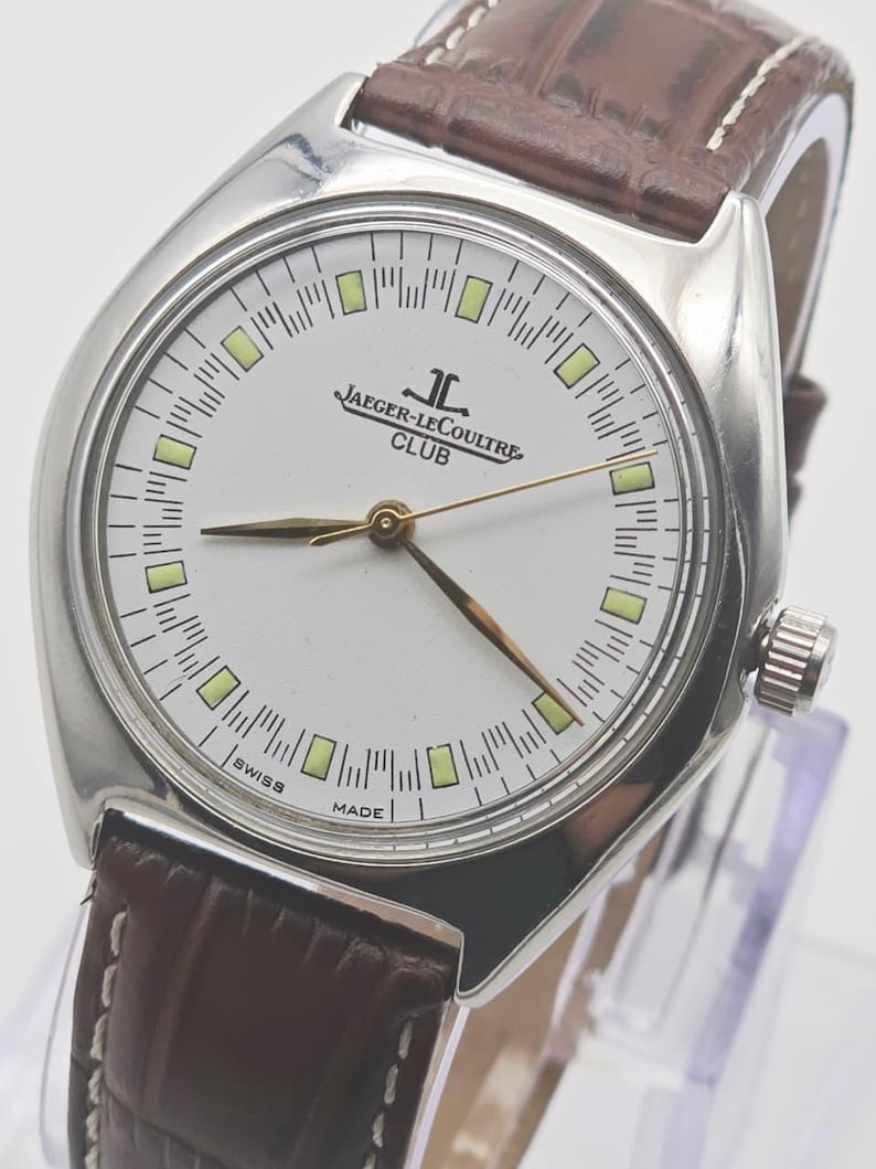 May include: A silver-toned Jaeger-LeCoultre Club watch with a white face, gold hands, and a brown leather strap. The watch face features the brand name and the word "Club". The watch has a classic design.