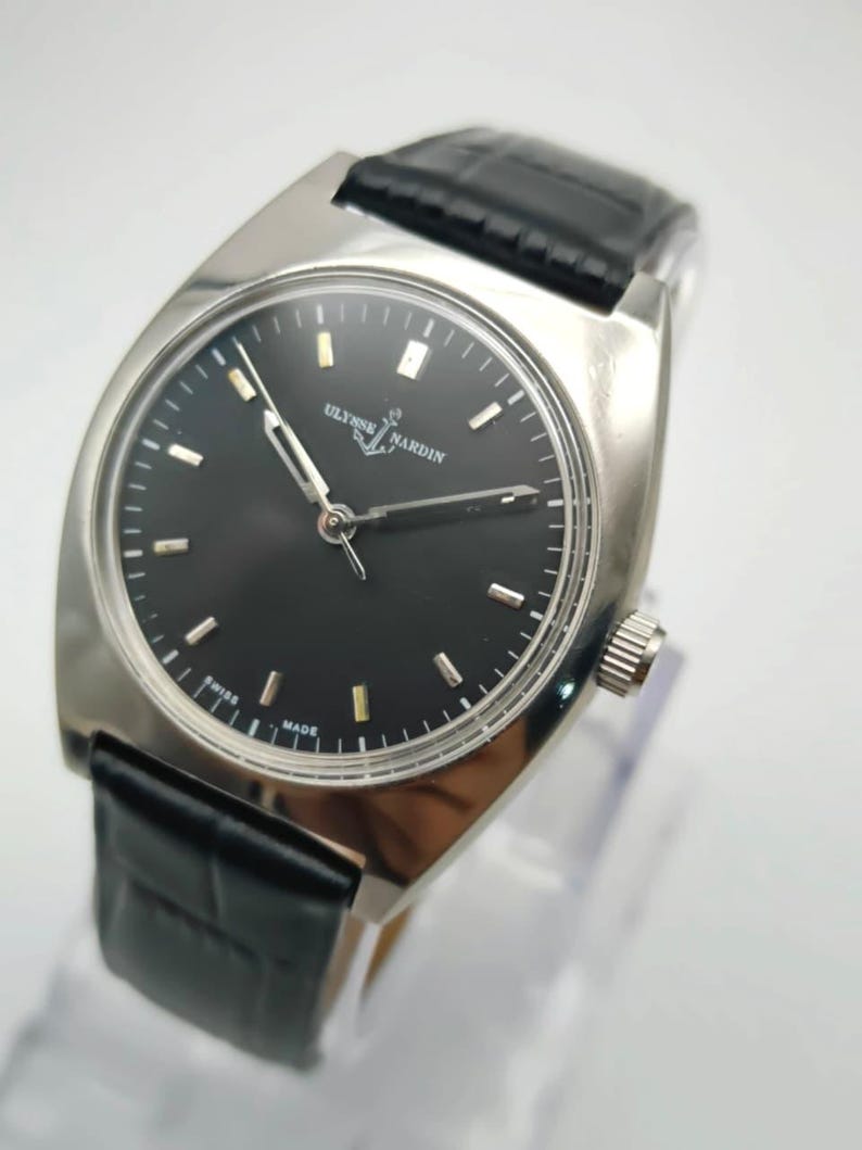 Puede incluir: Un reloj de pulsera plateado con esfera negra y correa de cuero negro. La esfera del reloj presenta marcadores de hora y agujas plateadas. La marca "ULYSSE NARDIN" se muestra en la esfera. El reloj es de dise&ntilde;o cl&aacute;sico.