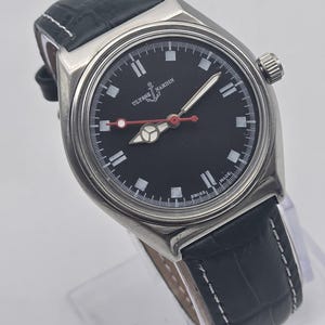 Puede incluir: Un reloj Ulysse Nardin de acero inoxidable con esfera negra y correa de cuero negro. El reloj presenta marcadores de hora blancos, un segundero rojo y una corona en el lado derecho. La correa mide unos 20 cm.
