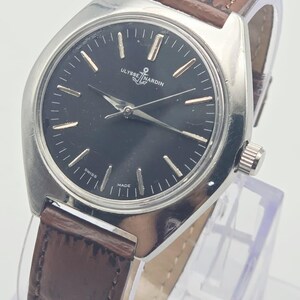 May include: A silver-toned Ulysse Nardin wristwatch with a black face and brown leather band. The watch face has gold-colored hour markers and hands. The band is a dark brown color. The watch is labeled "SWISS MADE".