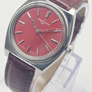 May include: A vintage Ulysse Nardin wristwatch with a rectangular stainless steel case and a burgundy leather strap. The watch features a red dial with silver-tone hour markers and hands. The watch face has the text "Ulysse Nardin" and "Swiss Made".