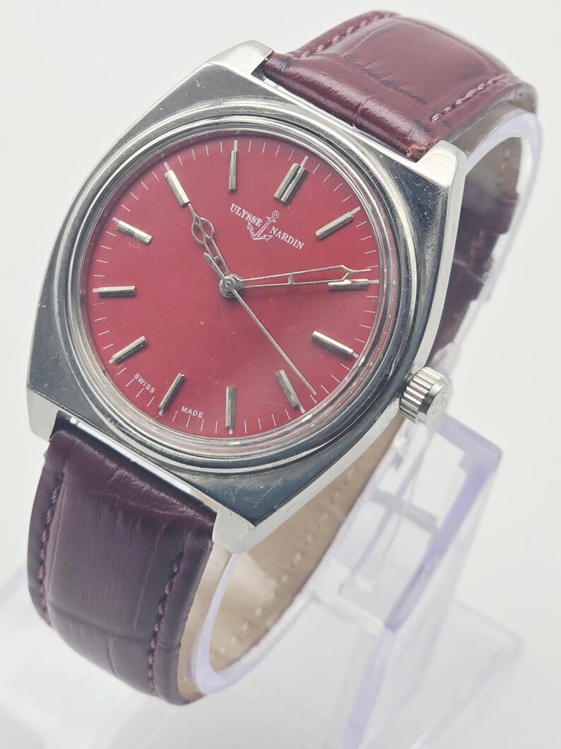 May include: A vintage Ulysse Nardin wristwatch with a rectangular stainless steel case and a burgundy leather strap. The watch features a red dial with silver-tone hour markers and hands. The watch face has the text "Ulysse Nardin" and "Swiss Made".