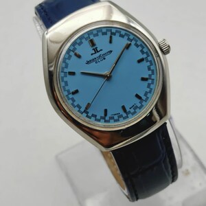 May include: A silver Jaeger-LeCoultre Club watch with a light blue face and a dark blue leather strap. The watch face has a patterned border and the brand name is displayed. The watch is a classic design.