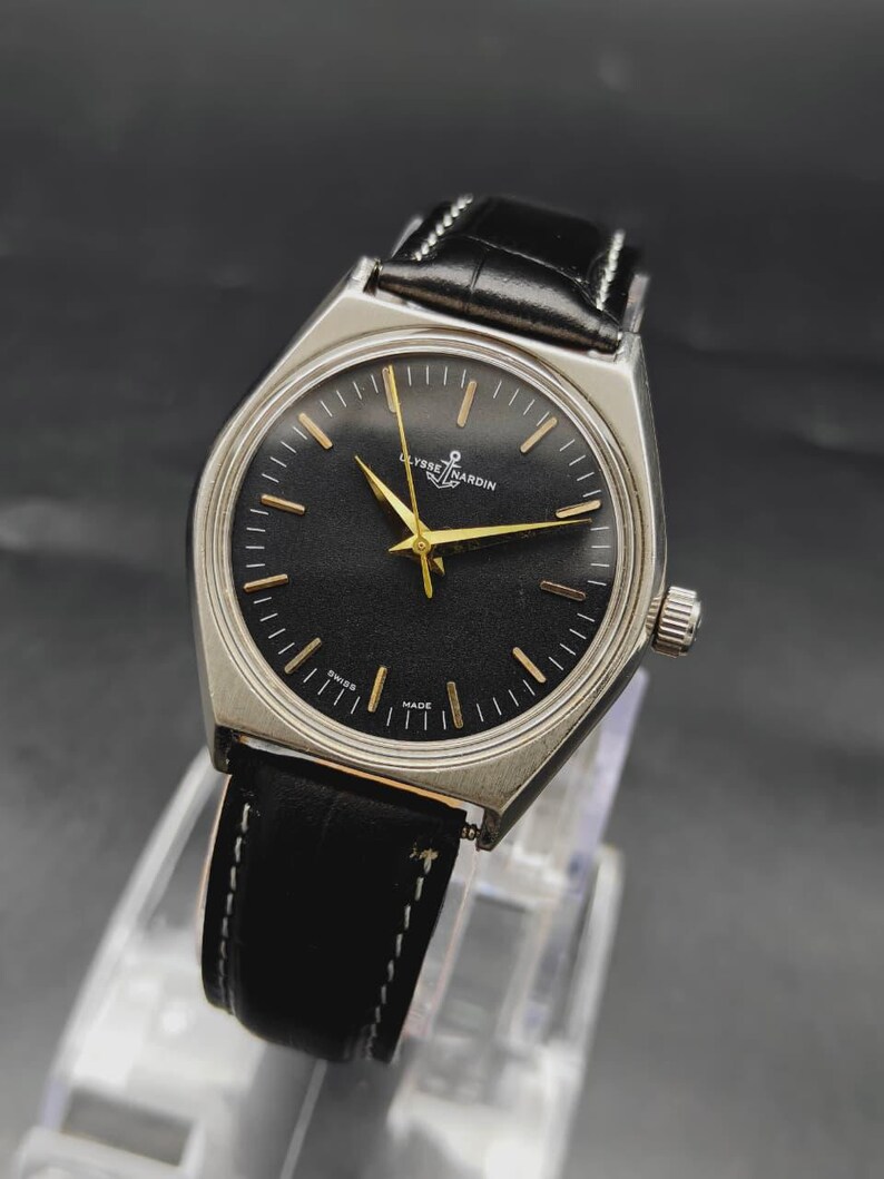 May include: A Ulysse Nardin wristwatch featuring a round black dial with gold-toned hour markers and hands. The watch has a silver-toned stainless steel case and a black leather strap with white stitching. The text Ulysse Nardin is printed on the dial.