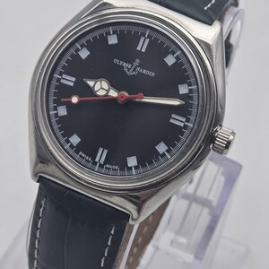 Puede incluir: Un reloj Ulysse Nardin de acero inoxidable con esfera negra y correa de cuero azul oscuro. La esfera presenta marcadores blancos, un segundero con punta roja y el nombre de la marca. La correa tiene costuras blancas.