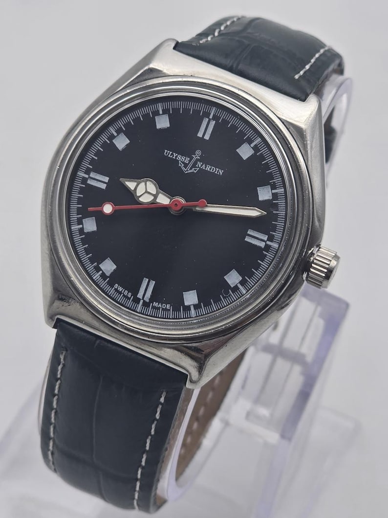 Puede incluir: Un reloj Ulysse Nardin de acero inoxidable con esfera negra y correa de cuero azul oscuro. La esfera presenta marcadores blancos, un segundero con punta roja y el nombre de la marca. La correa tiene costuras blancas.