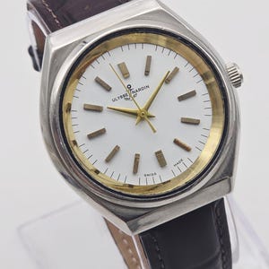 Pu&ograve; includere: Un orologio Ulysse Nardin argentato con quadrante bianco, dettagli dorati e cinturino in pelle marrone. L'orologio presenta lancette e indici dorati, con la scritta "SWISS MADE". L'orologio ha una forma rettangolare.