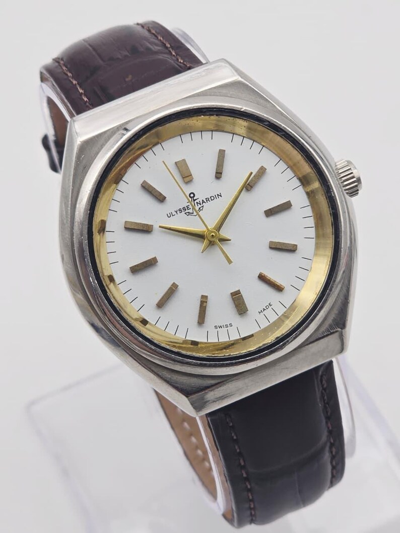 Pu&ograve; includere: Un orologio Ulysse Nardin argentato con quadrante bianco, dettagli dorati e cinturino in pelle marrone. L'orologio presenta lancette e indici dorati, con la scritta "SWISS MADE". L'orologio ha una forma rettangolare.
