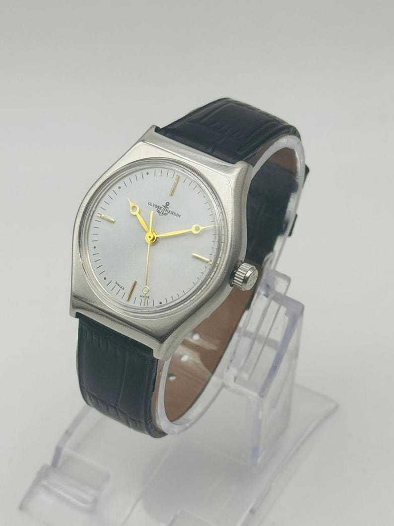 May include: A silver-toned wristwatch with a white face and gold hands. The watch has a black leather band and a small crown. The watch is displayed on a clear stand.