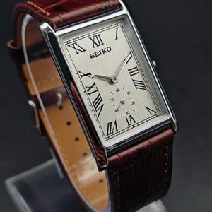 May include: A rectangular Seiko watch with a silver face, black Roman numerals, and a small seconds dial. The watch has a brown leather band. The watch case is silver-toned.
